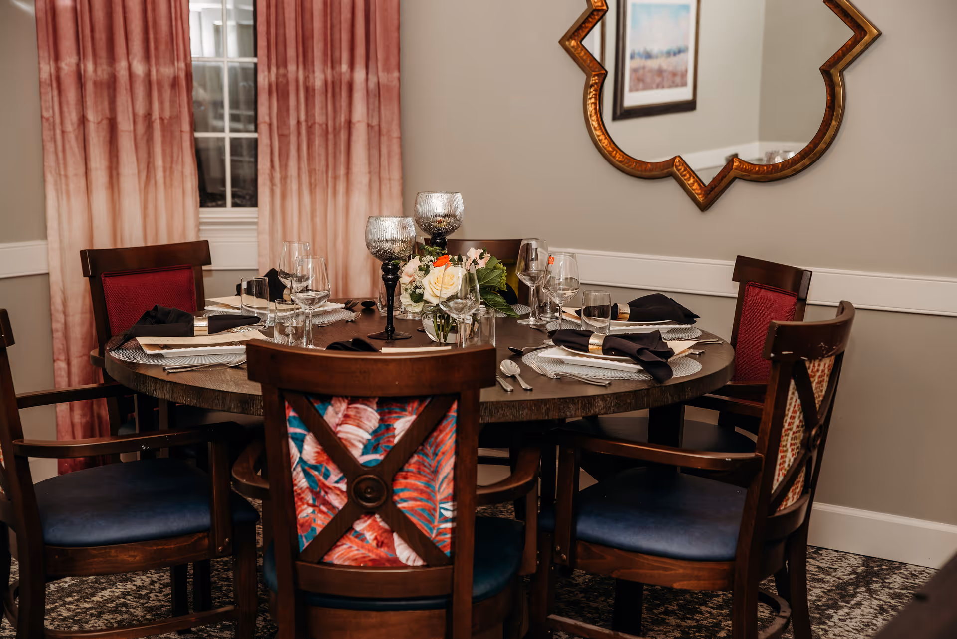 A round dining table set for six with dark wooden chairs featuring colorful patterned backs and blue cushions. The table is elegantly arranged with placemats, black napkins, glassware, silverware, and decorative candle holders. A floral centerpiece is placed in the middle. Behind the table, there are pink ombre curtains covering a window and a decorative mirror on the wall reflecting part of a framed picture.