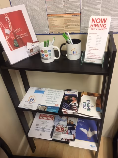 A dark, two-shelf brochure stand with pamphlets, mugs, and a Now Hiring sign indoors.