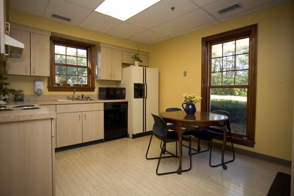 Bright kitchen with cabinets, fridge, microwave, and a round table by large windows.