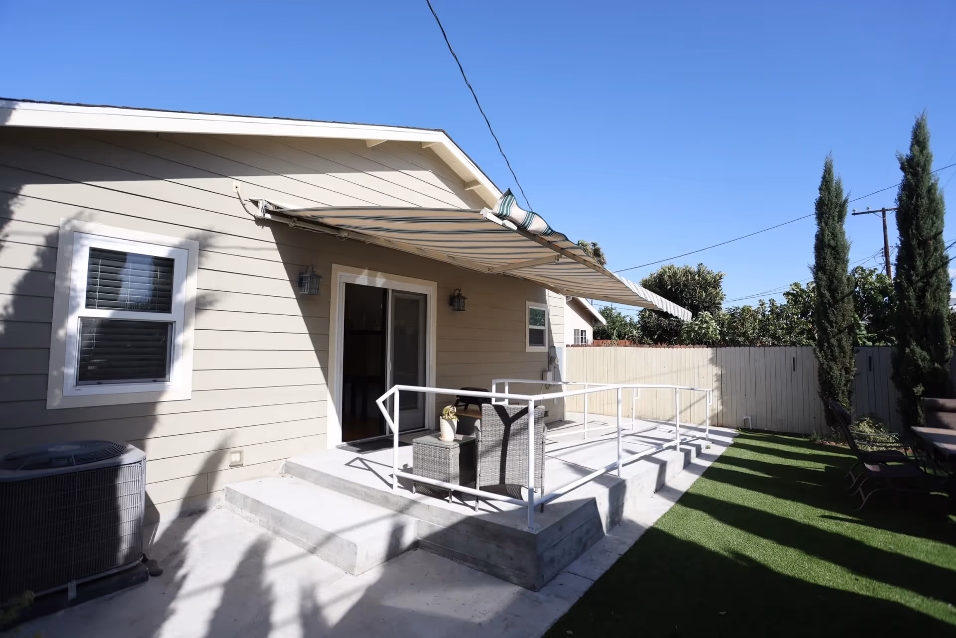 Backyard patio with a wheelchair-accessible ramp, outdoor seating, and a striped retractable awning on a beige single-story house.