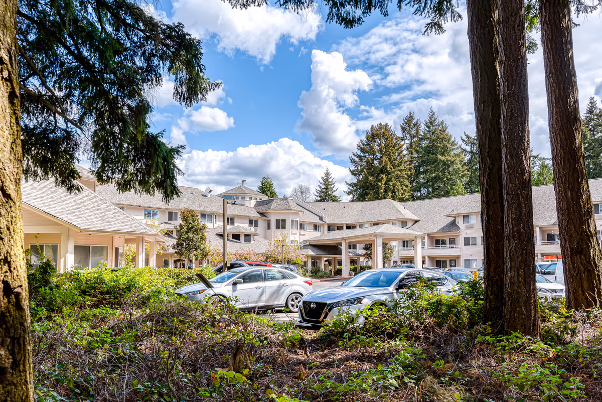 Front exterior of a senior living building with parked cars, trees, and a blue sky in the background.