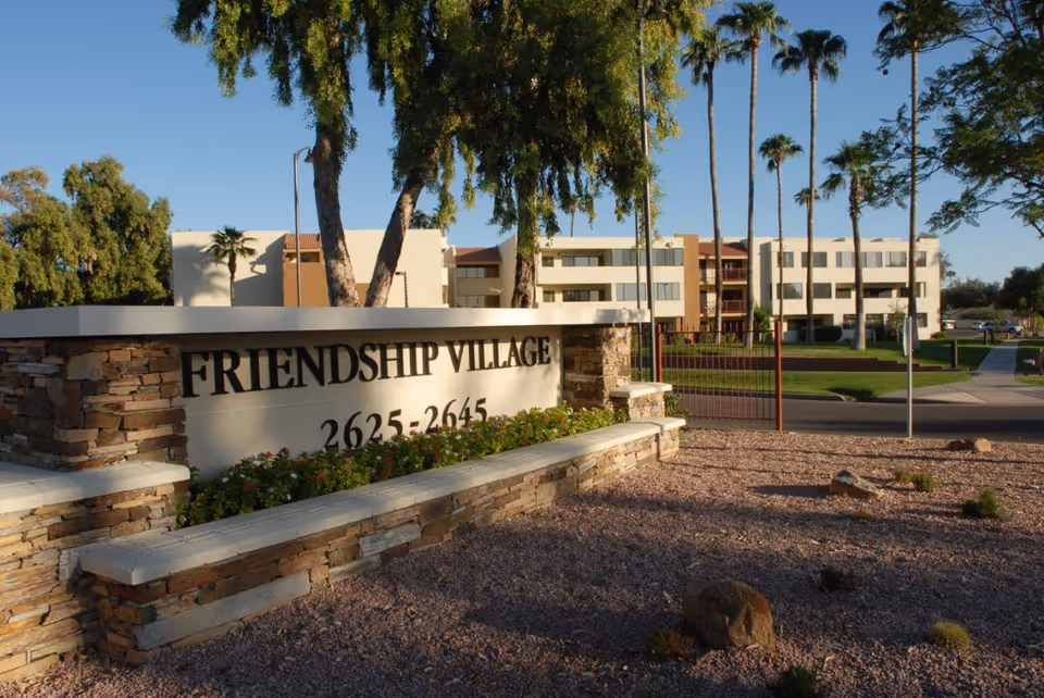 Stone entrance sign reading 'FRIENDSHIP VILLAGE' with multi-story residential buildings and palm trees in the background.