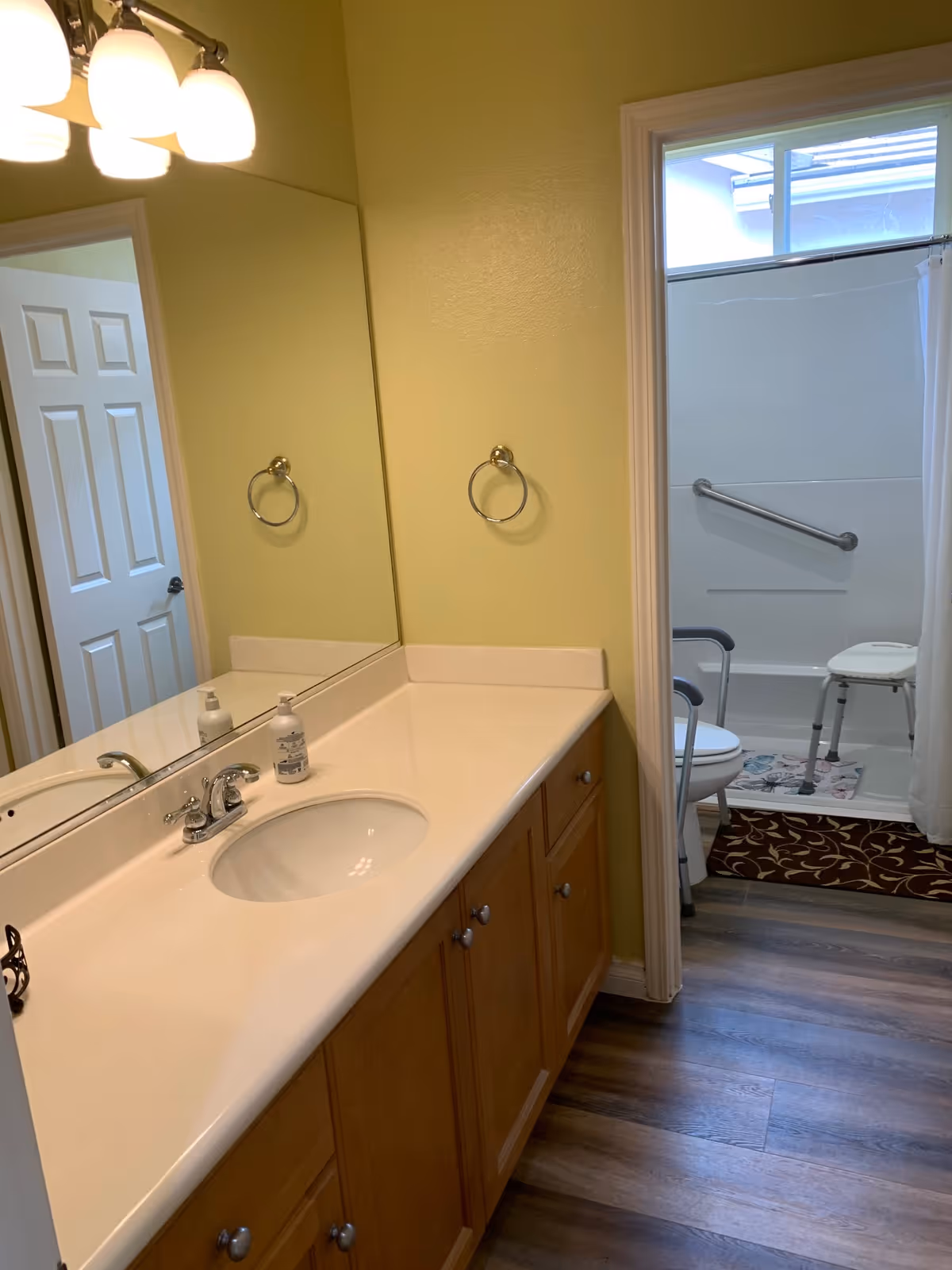 A bathroom with a long countertop featuring a sink and faucet, a large mirror above, and a light fixture with three bulbs. The walls are painted light yellow. In the background, there is a shower area with a grab bar, a shower chair, and a toilet partially visible. The floor has wood-like vinyl flooring.