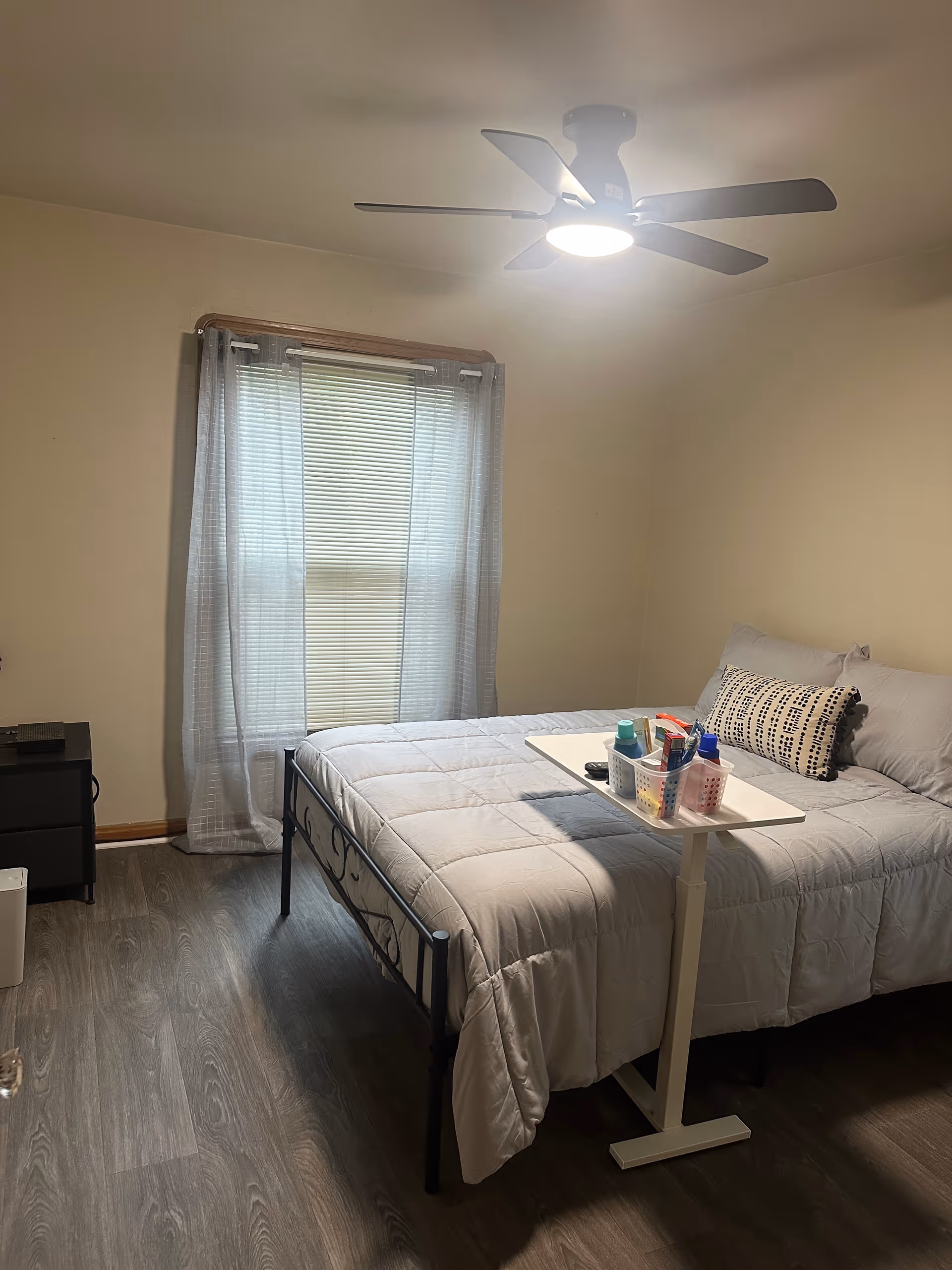 Cozy bedroom with a bed, ceiling fan, window blinds, and a small side table.