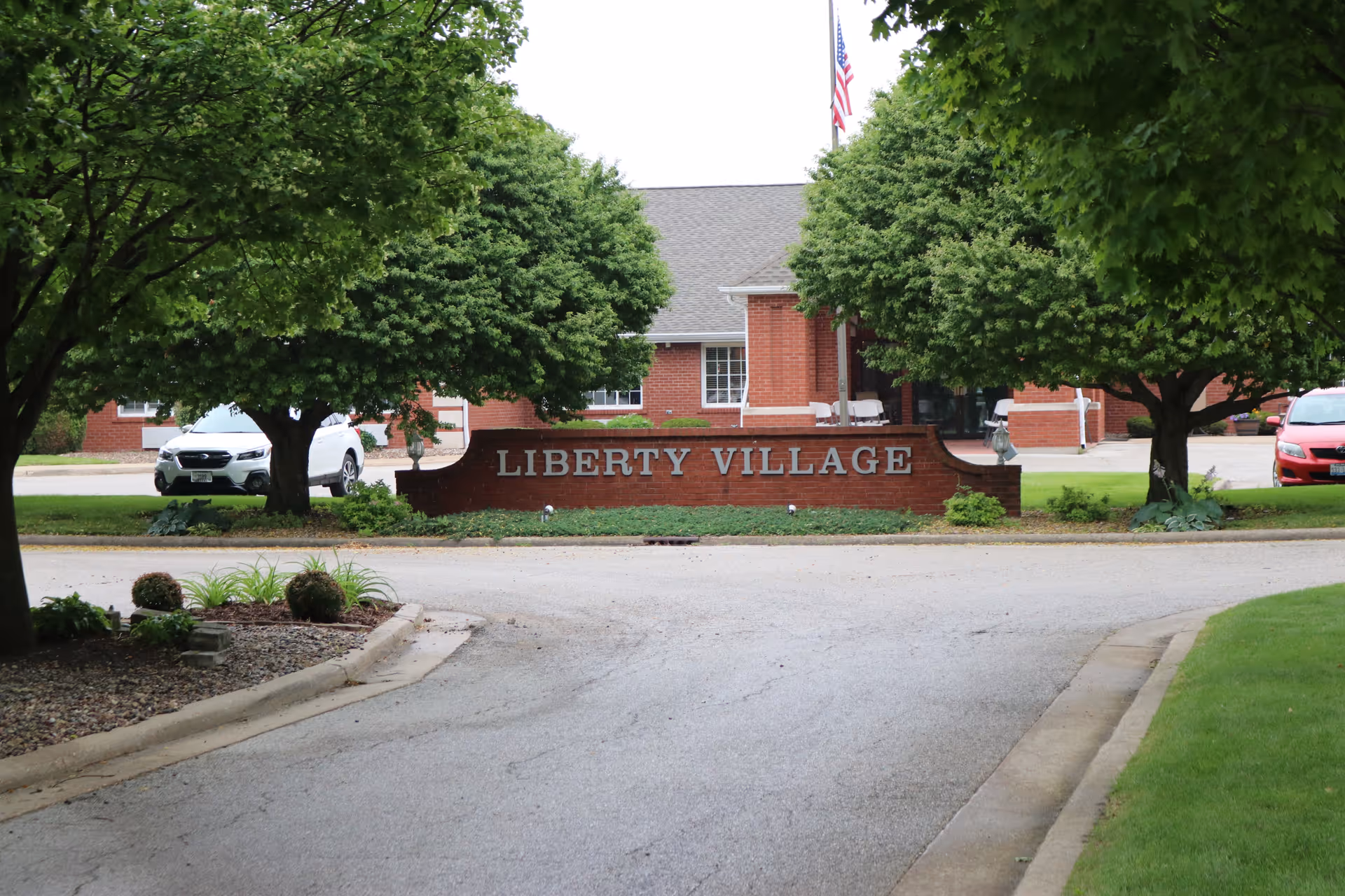 Brick entrance sign reading "LIBERTY VILLAGE" at a driveway entrance with trees and the facility building visible behind it.