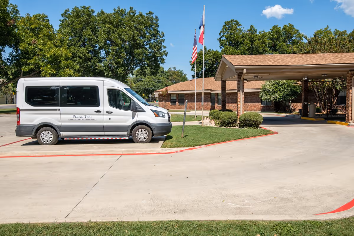 Exterior view of Pecan Tree Rehab and Healthcare Center showing a white van parked in front of a brick building with a covered entrance. Two flagpoles with the American and Texas flags are visible near the entrance, surrounded by green trees and bushes under a clear blue sky.