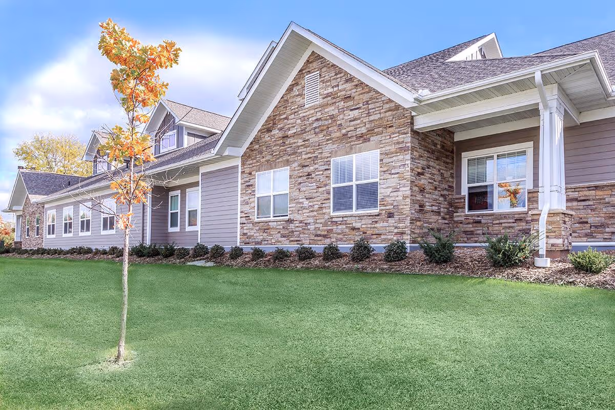 Exterior view of a single-story assisted living facility building with stone and siding walls, multiple windows, a small tree with autumn leaves in the foreground, and a well-maintained green lawn under a partly cloudy sky.