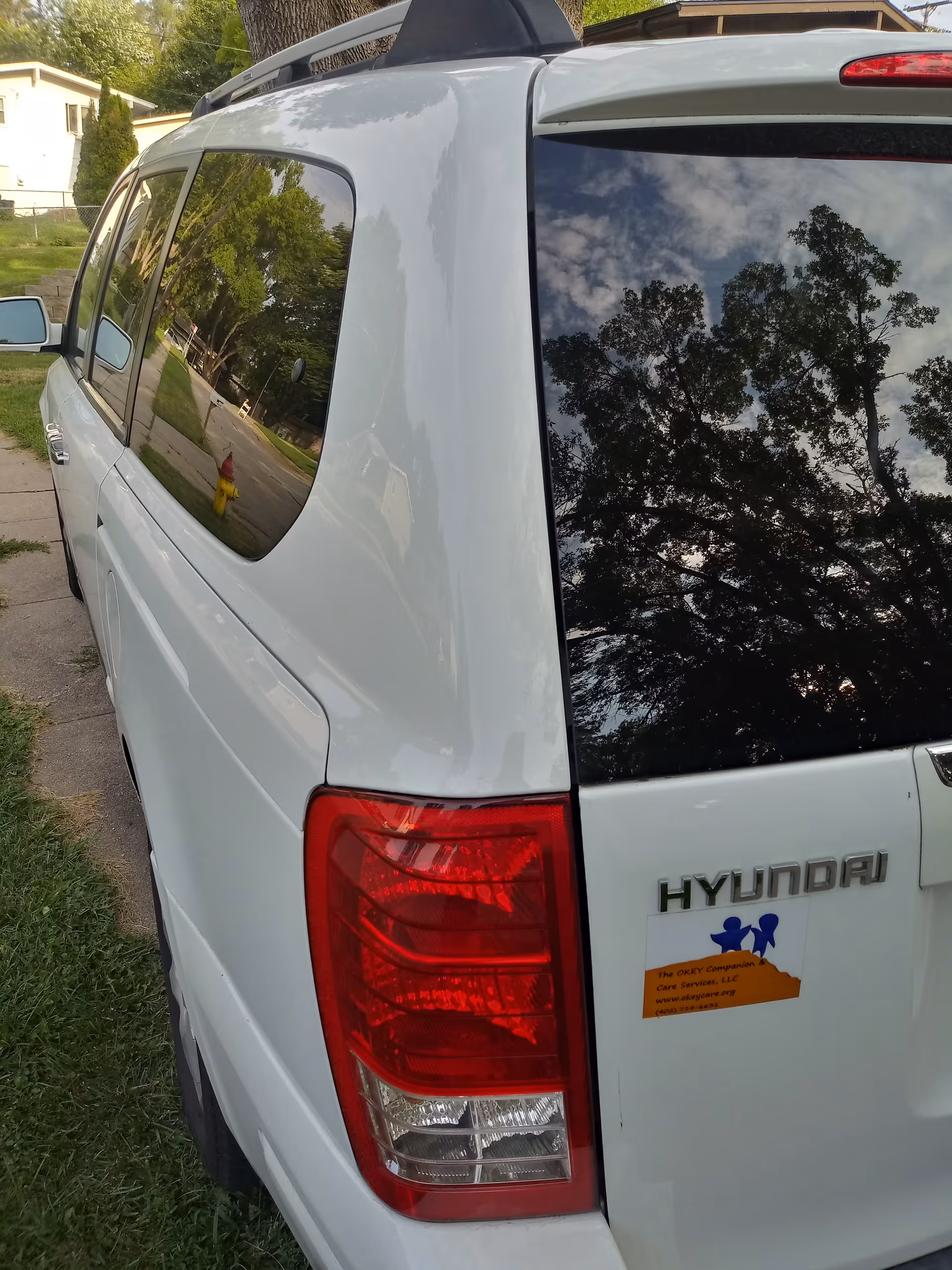 Rear of a white Hyundai van parked on a residential street, reflecting trees in windows.