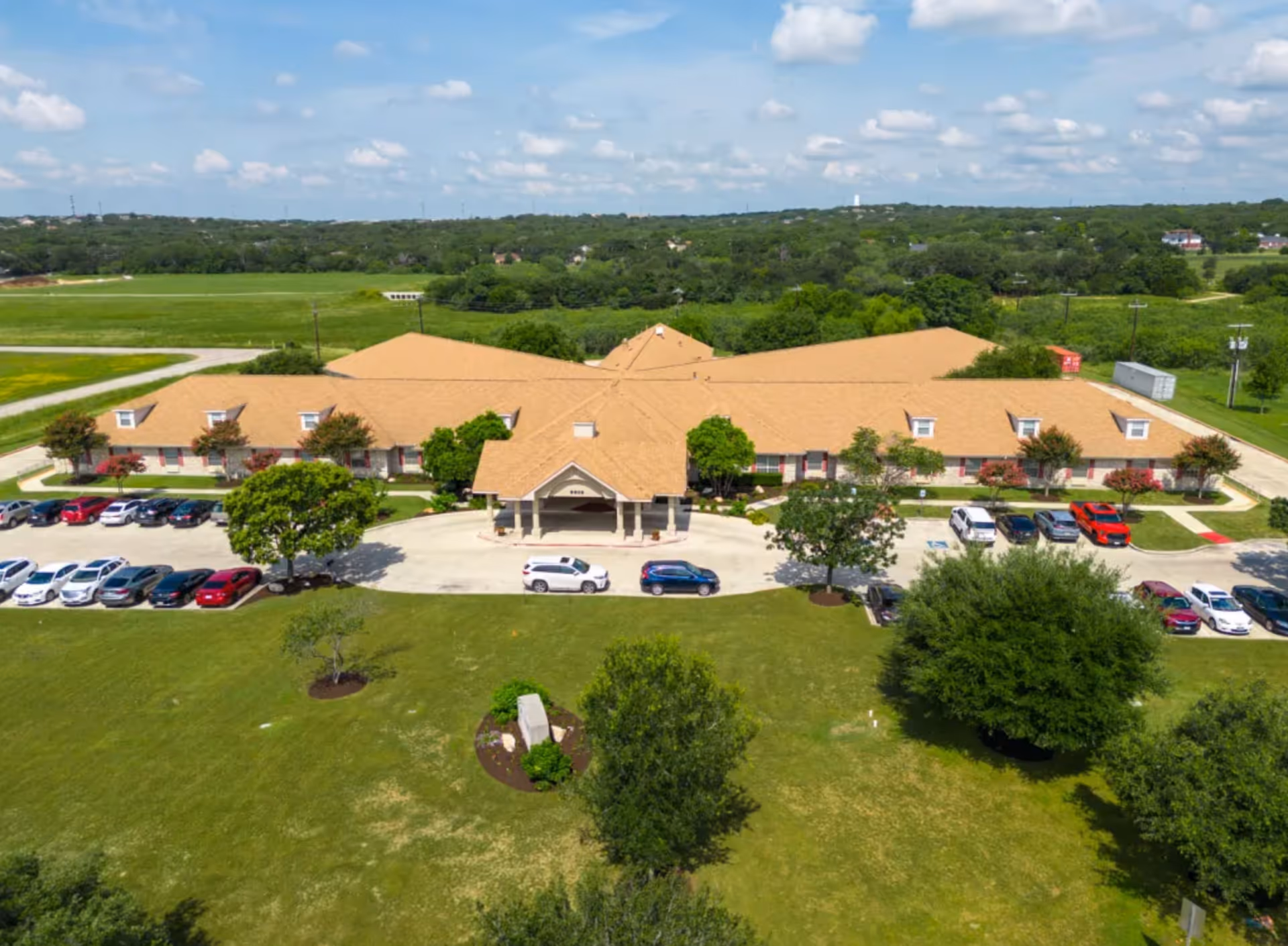 Aerial view of Mystic Park Nursing and Rehabilitation Center, a single-story building with a tan roof surrounded by green lawns, trees, and a parking lot with several cars. The sky is partly cloudy.