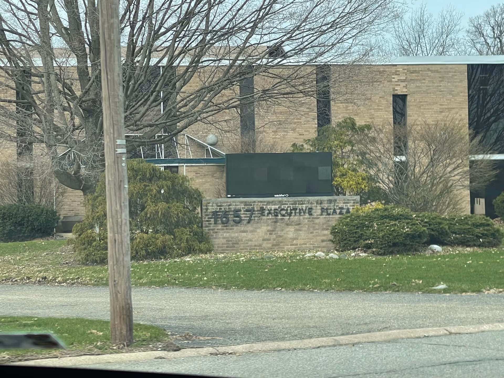 Brick facility exterior with a brick wall sign, trees, shrubs, and a street in foreground.