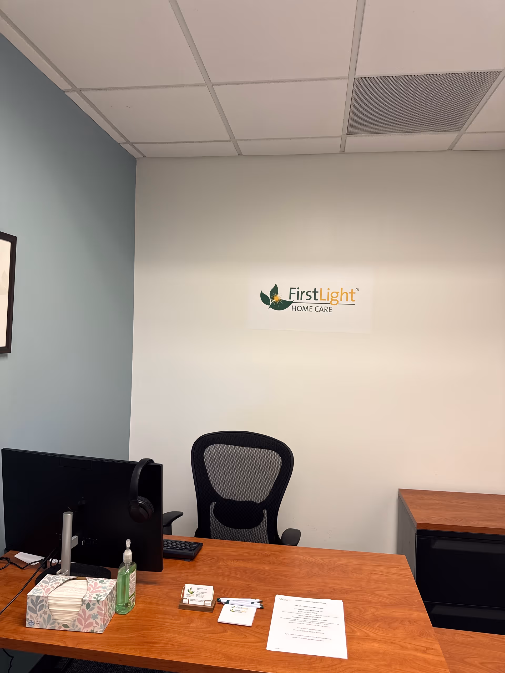 Office desk with computer, chair, tissue box, and FirstLight Home Care sign on wall.