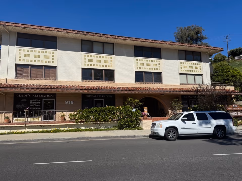 Two-story beige building with storefronts on the ground floor and a white SUV parked nearby.