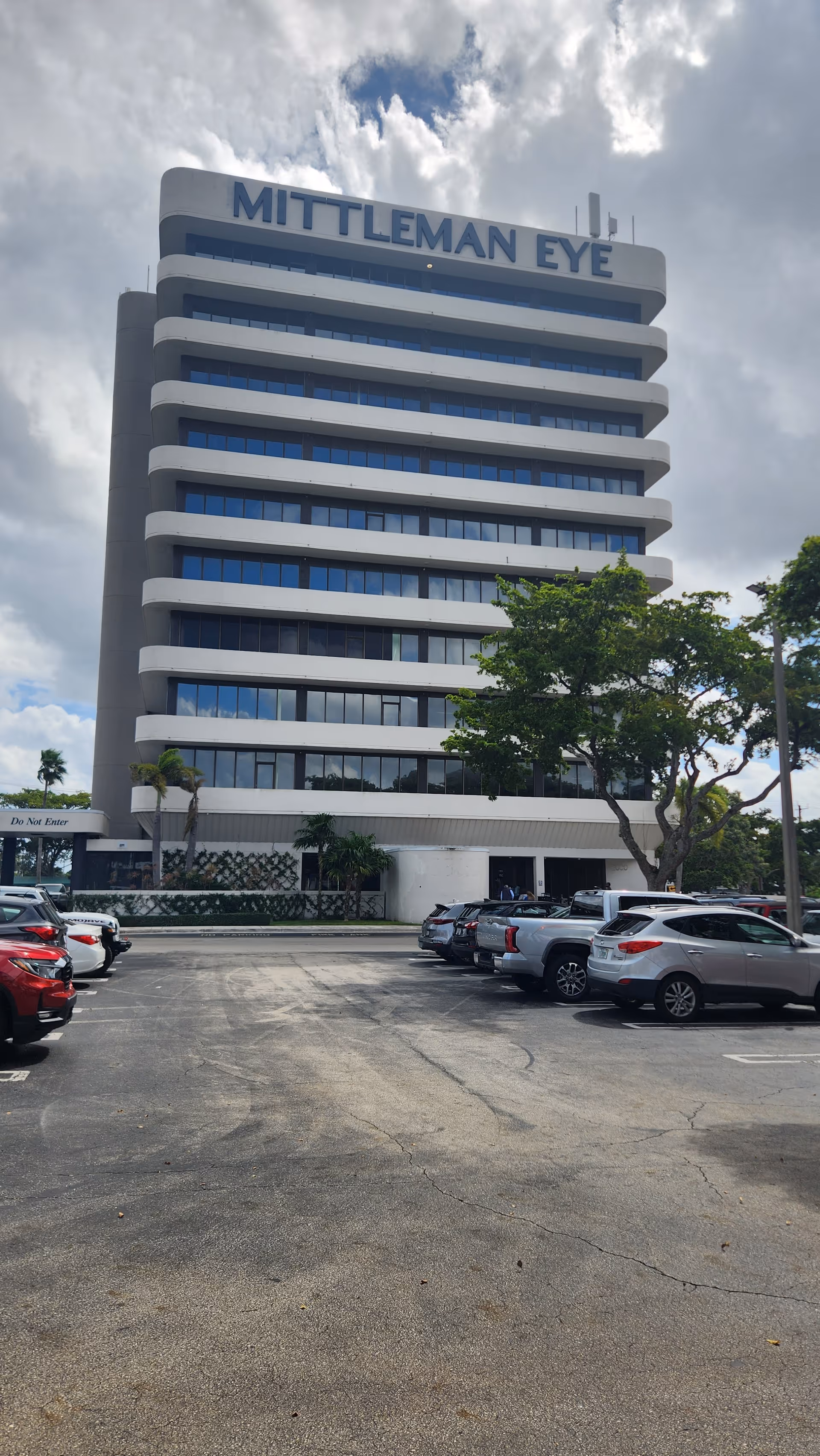 Tall curved white office building labeled Mittleman Eye beside a parking lot.