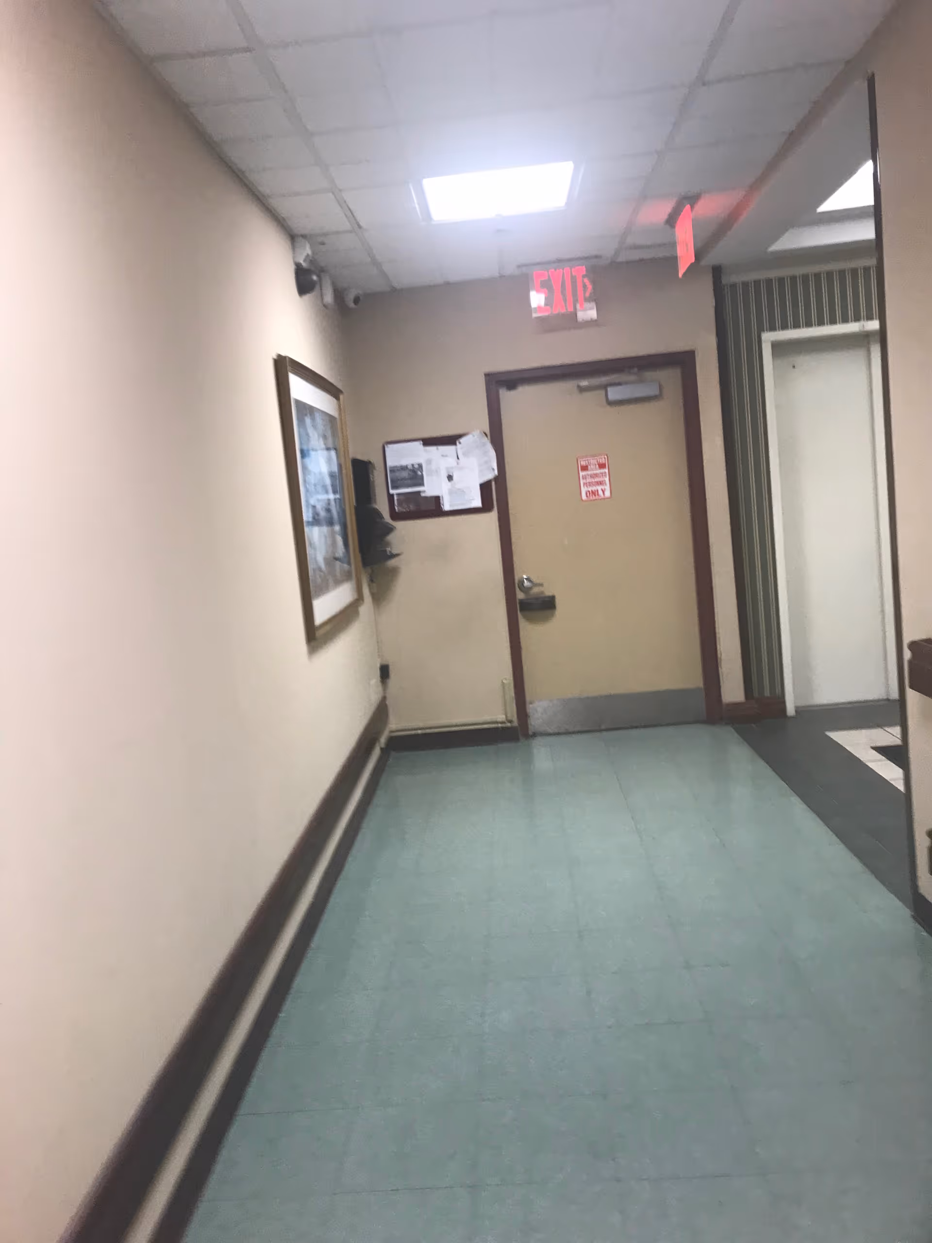 Hallway inside a senior living facility with a door, exit sign, bulletin board, and photo.