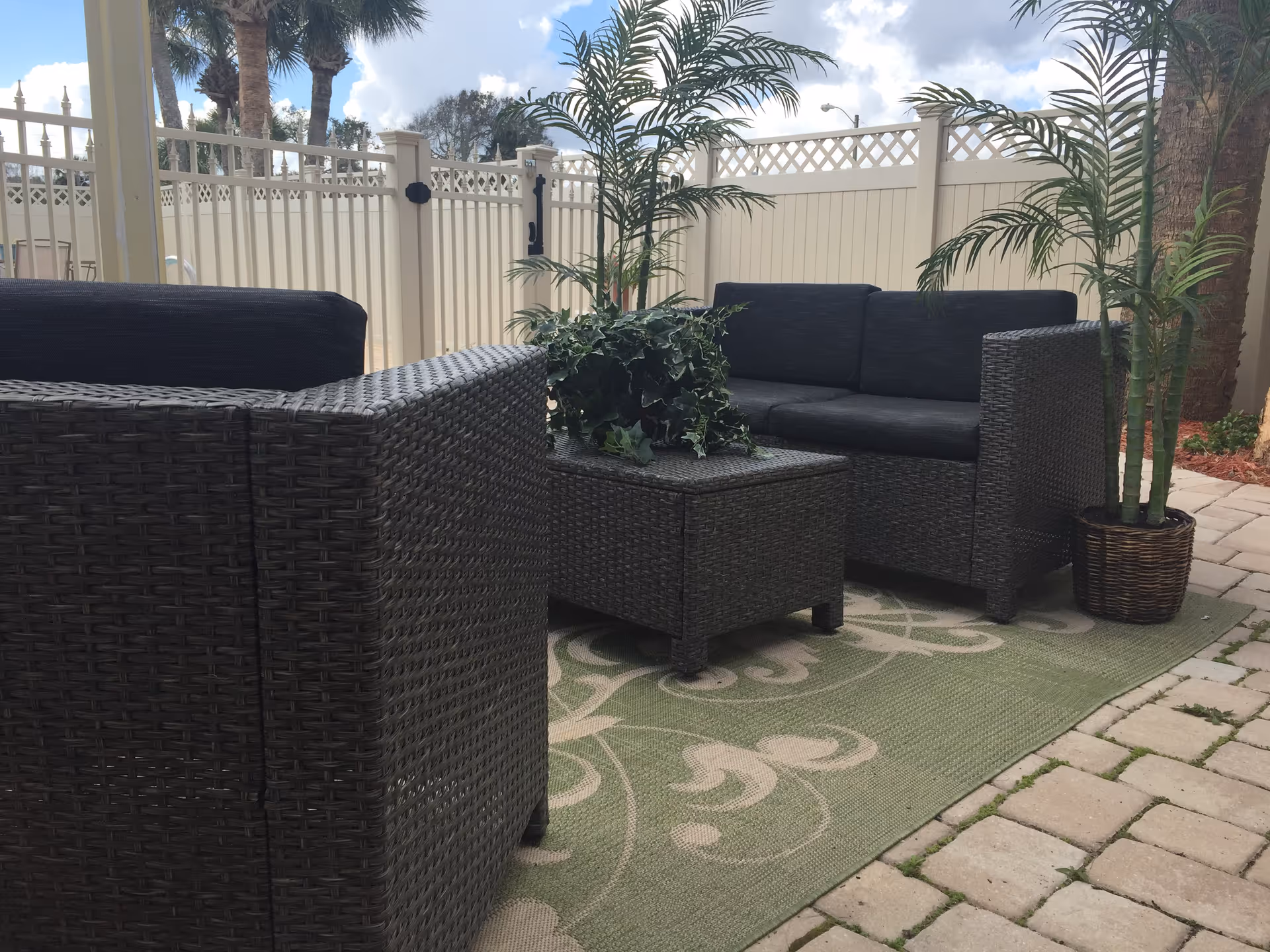 Outdoor patio with wicker furniture, potted plants, and a patterned rug near a fence.