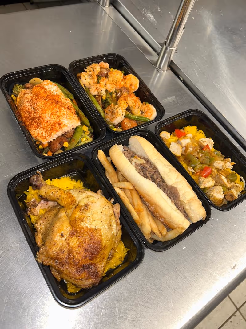 Five black meal trays with assorted cooked dishes on stainless steel counter.