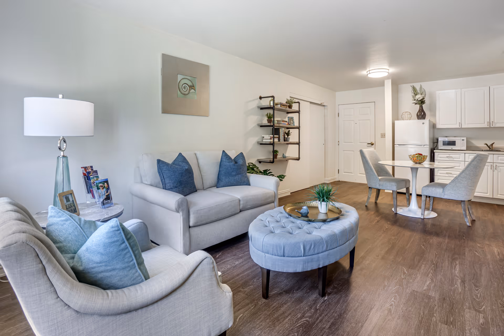 A bright and modern living area in a senior living facility featuring a light gray sofa with blue pillows, a matching armchair with blue cushions, a round tufted ottoman with a decorative tray and plant, a small round dining table with two gray upholstered chairs, and a kitchenette with white cabinets, a refrigerator, microwave, and sink. The walls are white with a framed shell artwork and a wall-mounted shelf holding books and plants.