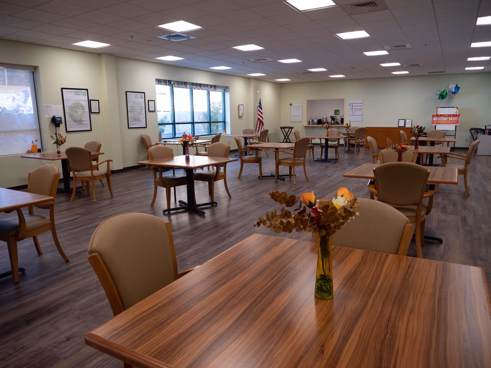 A spacious senior dining room with multiple tables, chairs, flowers, and an American flag in background.