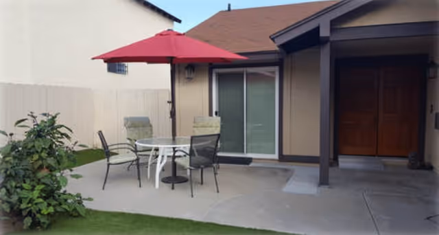 Small backyard patio with a round table, four chairs and a red umbrella in front of a sliding glass door and entryway.