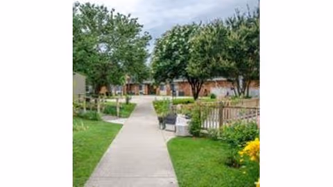A paved walkway through a garden area with green grass, trees, and bushes on both sides. There are benches along the path and a building with a brick exterior in the background under a partly cloudy sky.