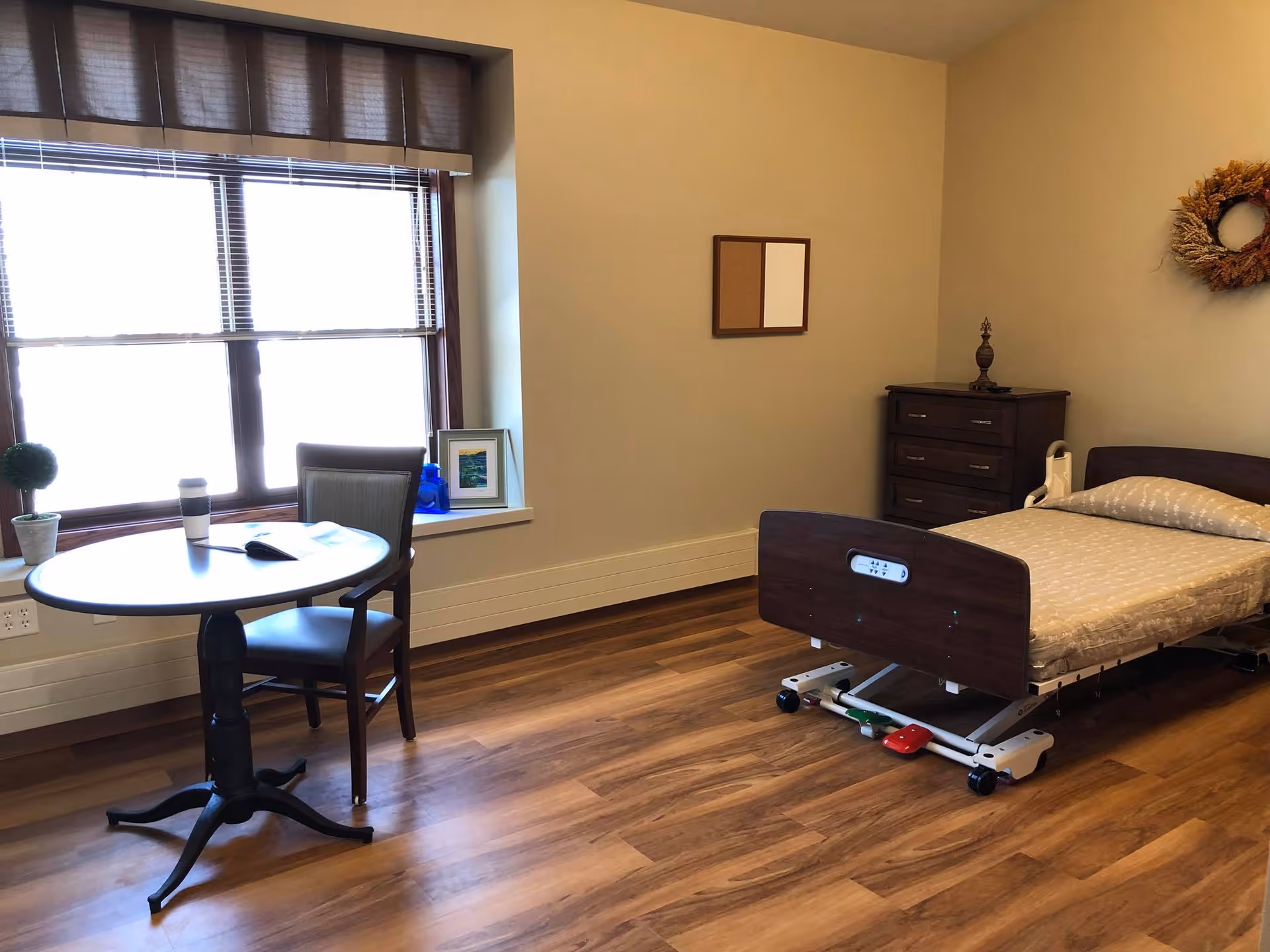 A simple, clean assisted living bedroom with a hospital-style adjustable bed, a wooden dresser, a small round table with a chair, a window with blinds, and minimal decorations including a small plant, a framed picture, and a wreath on the wall.