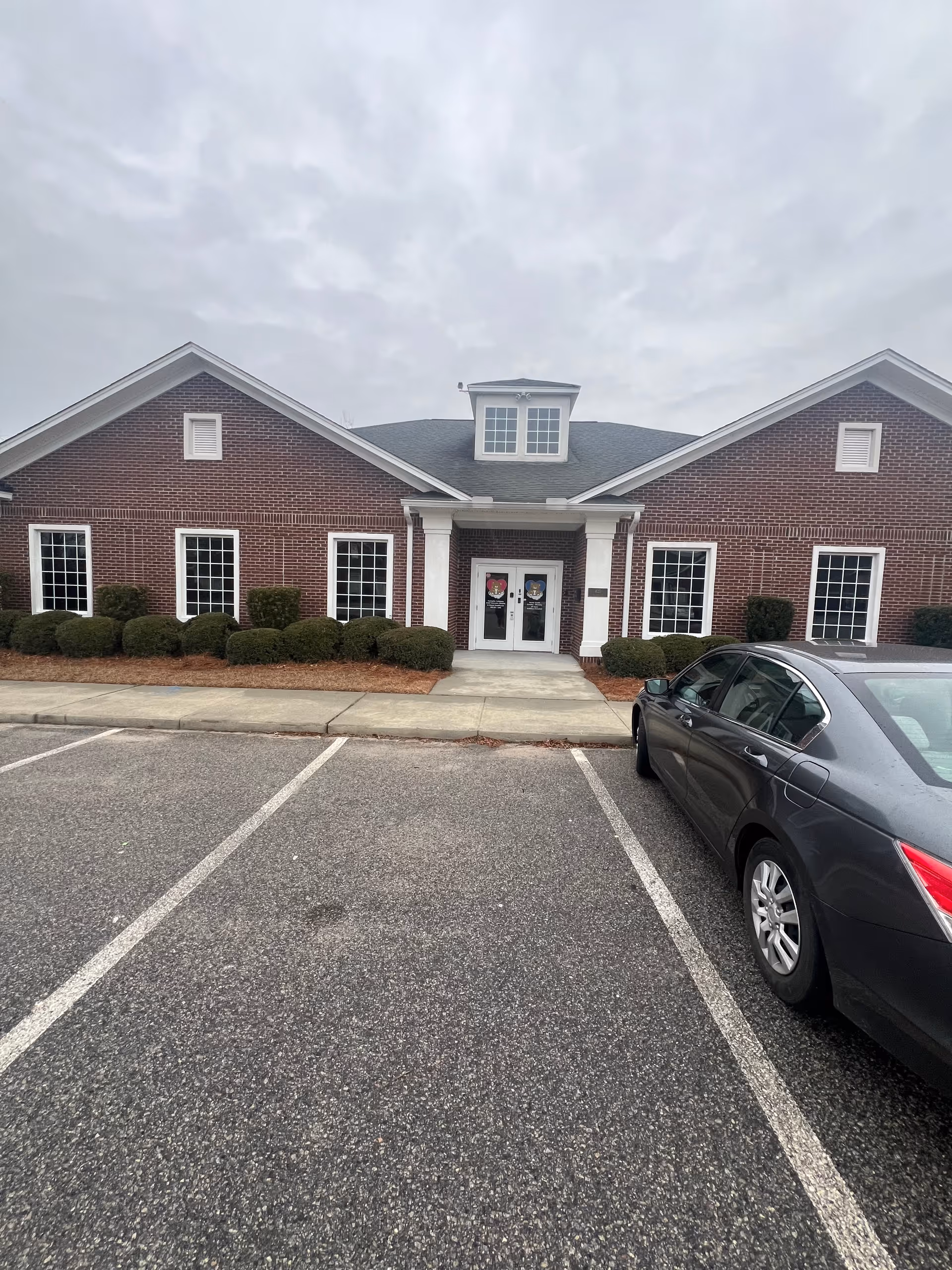 Brick building with white columns and double doors, shrubs along entry, and a parked sedan.