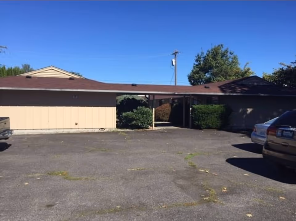 Single-story beige building with a carport and parked cars in a paved lot.