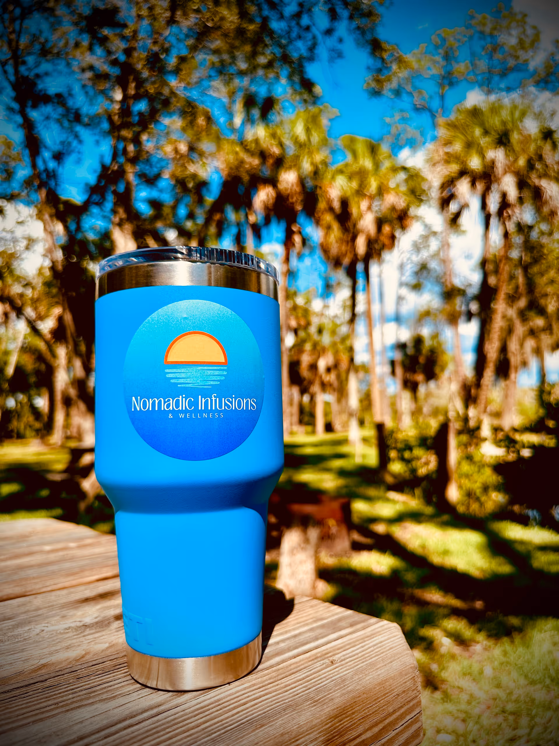 Blue Nomadic Infusions tumbler with logo on a wooden table outdoors.