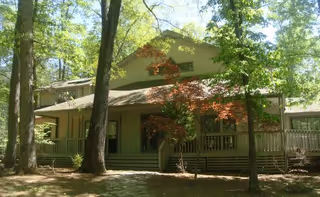 A large house surrounded by tall trees with green leaves. The house has a wide porch with steps leading up to it and a sloped roof. Sunlight filters through the trees, casting shadows on the ground and the house.