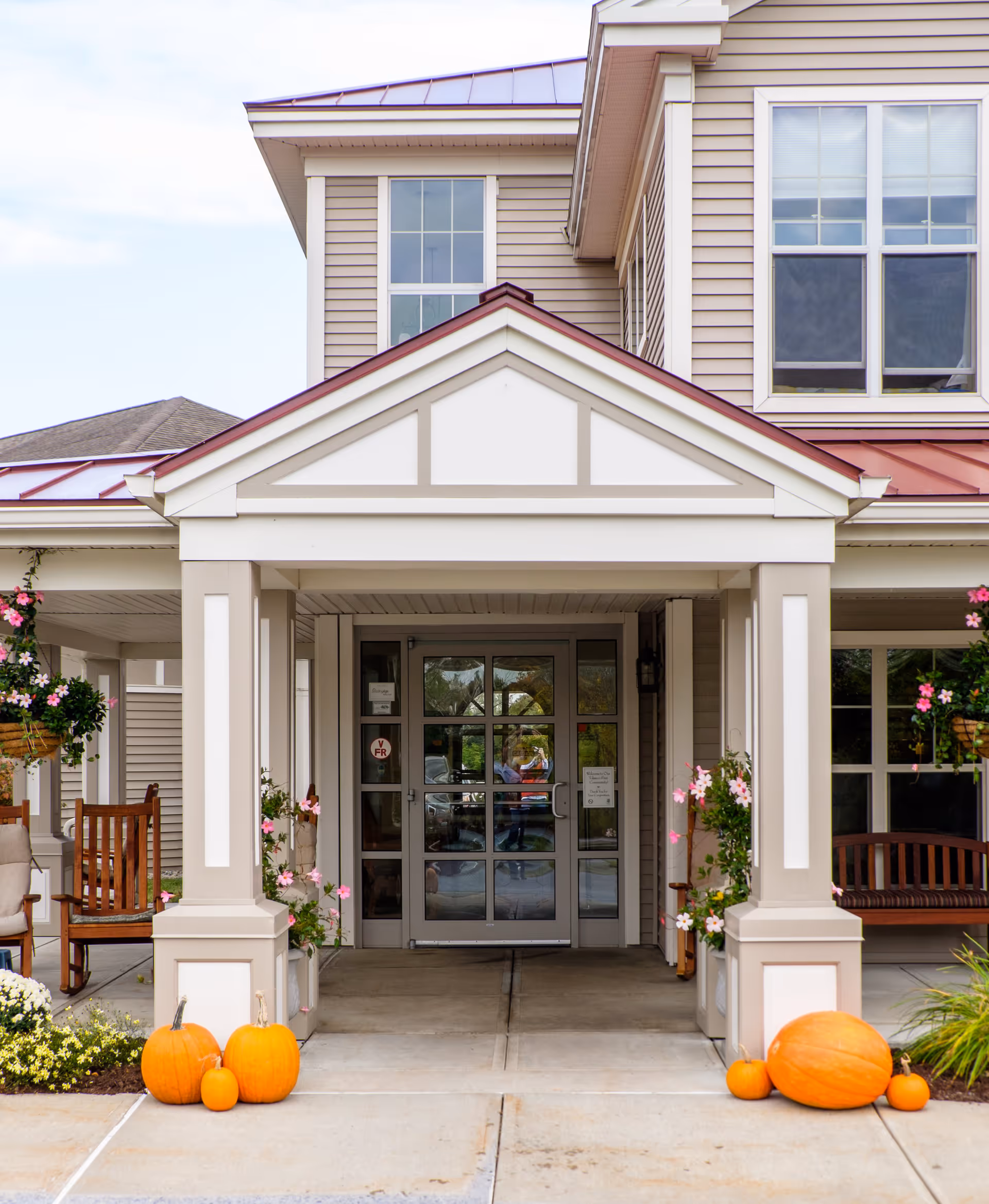 Entrance of a senior living facility with a covered porch supported by white columns. There are pumpkins and flowers decorating the entrance, wooden chairs and benches on either side, and large windows on the building facade.