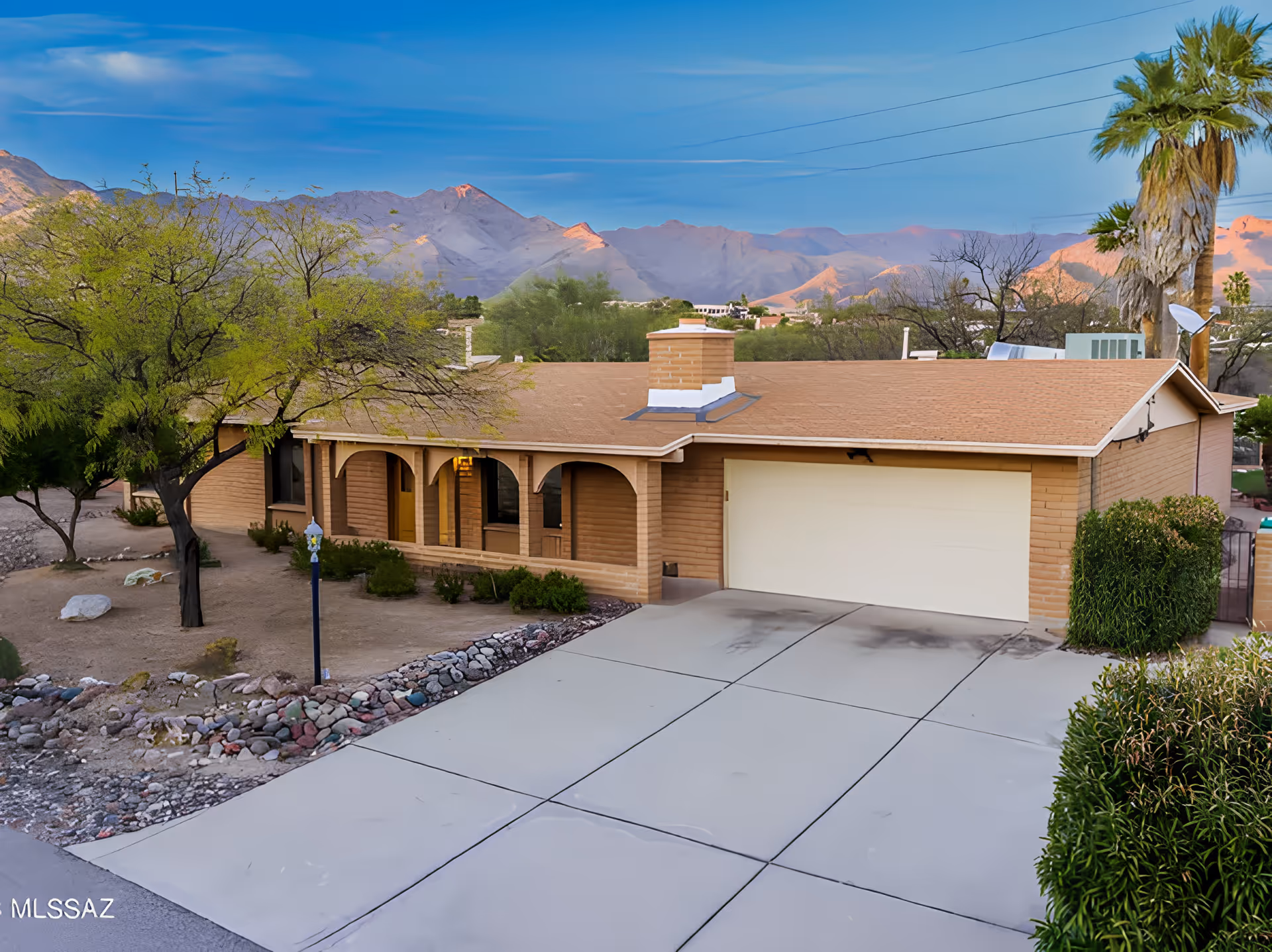 Single-story house with a beige exterior, a two-car garage, and a covered front porch with arches. The driveway is concrete and there are desert landscaping elements including rocks, bushes, and trees. Mountains are visible in the background under a clear blue sky.