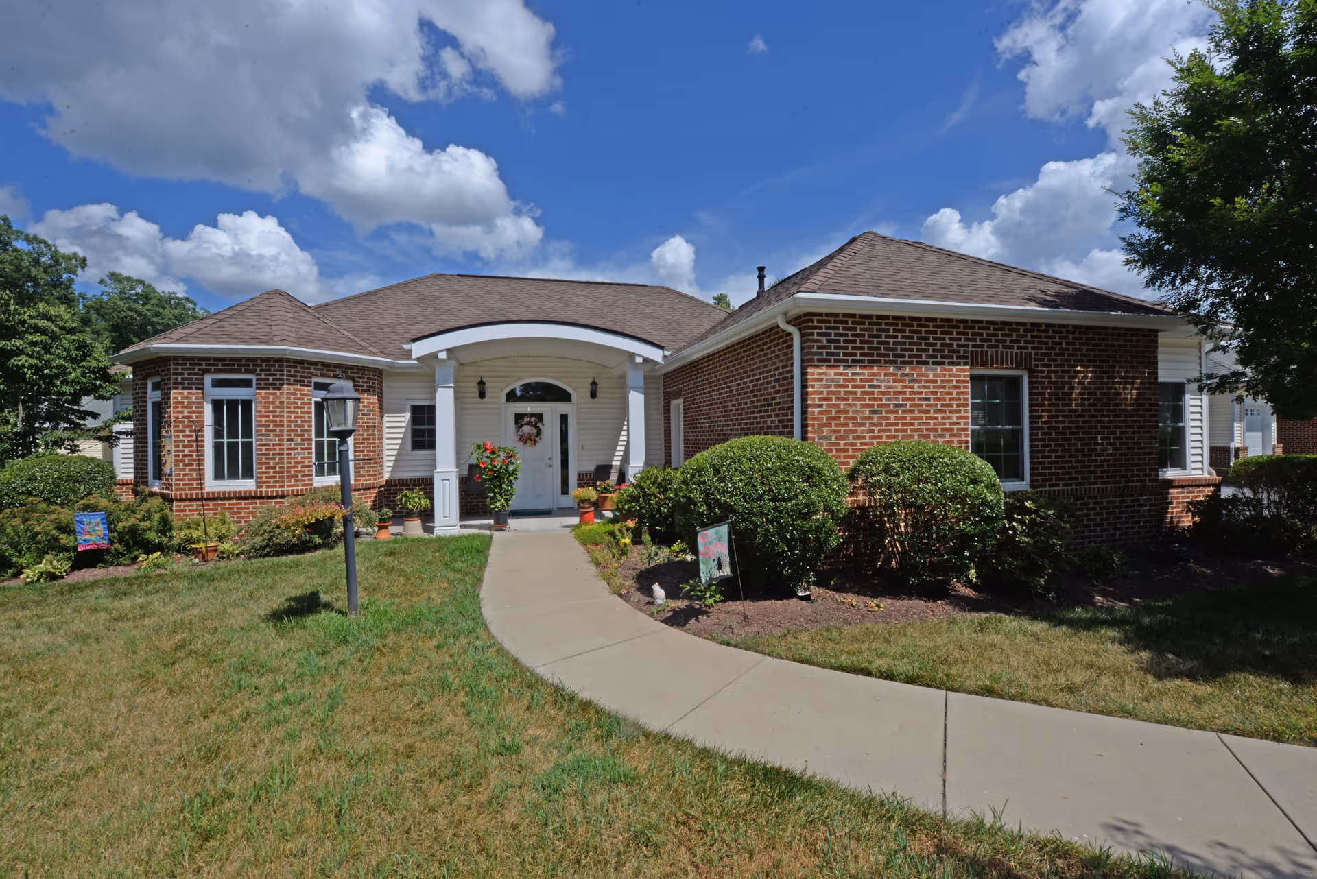 Front exterior view of a single-story brick and siding building with a curved walkway leading to a white entrance door under a small portico. The building is surrounded by green grass, bushes, and small plants under a partly cloudy blue sky.