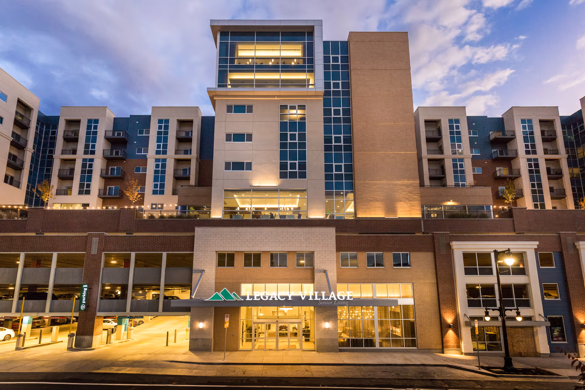 Exterior front view of Legacy Village of Sugar House building at dusk, showing a multi-story senior living facility with large windows, balconies, and an entrance with illuminated signage.