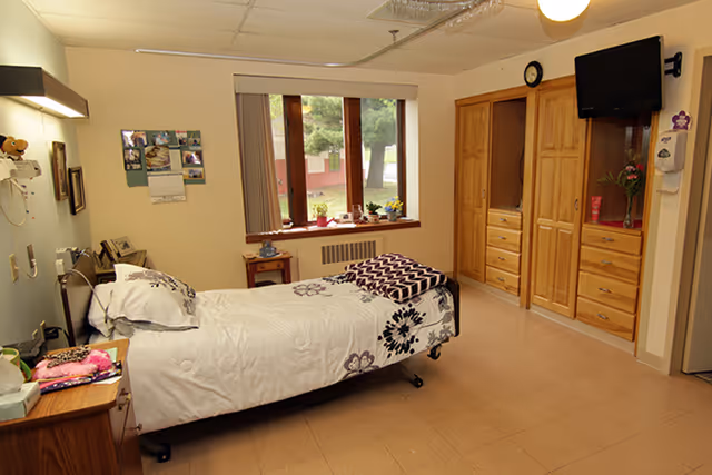 A single bed with white and floral bedding in a senior living facility room. The room has a window with a view of trees and a building outside. There is a wooden dresser with multiple drawers and a mounted TV on the wall. A small table with plants and personal items is placed near the window. The floor is tiled, and the walls are painted light colors.