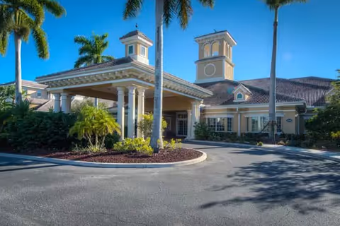 Exterior view of a senior living facility building with a covered entrance, palm trees, and landscaping under a clear blue sky.