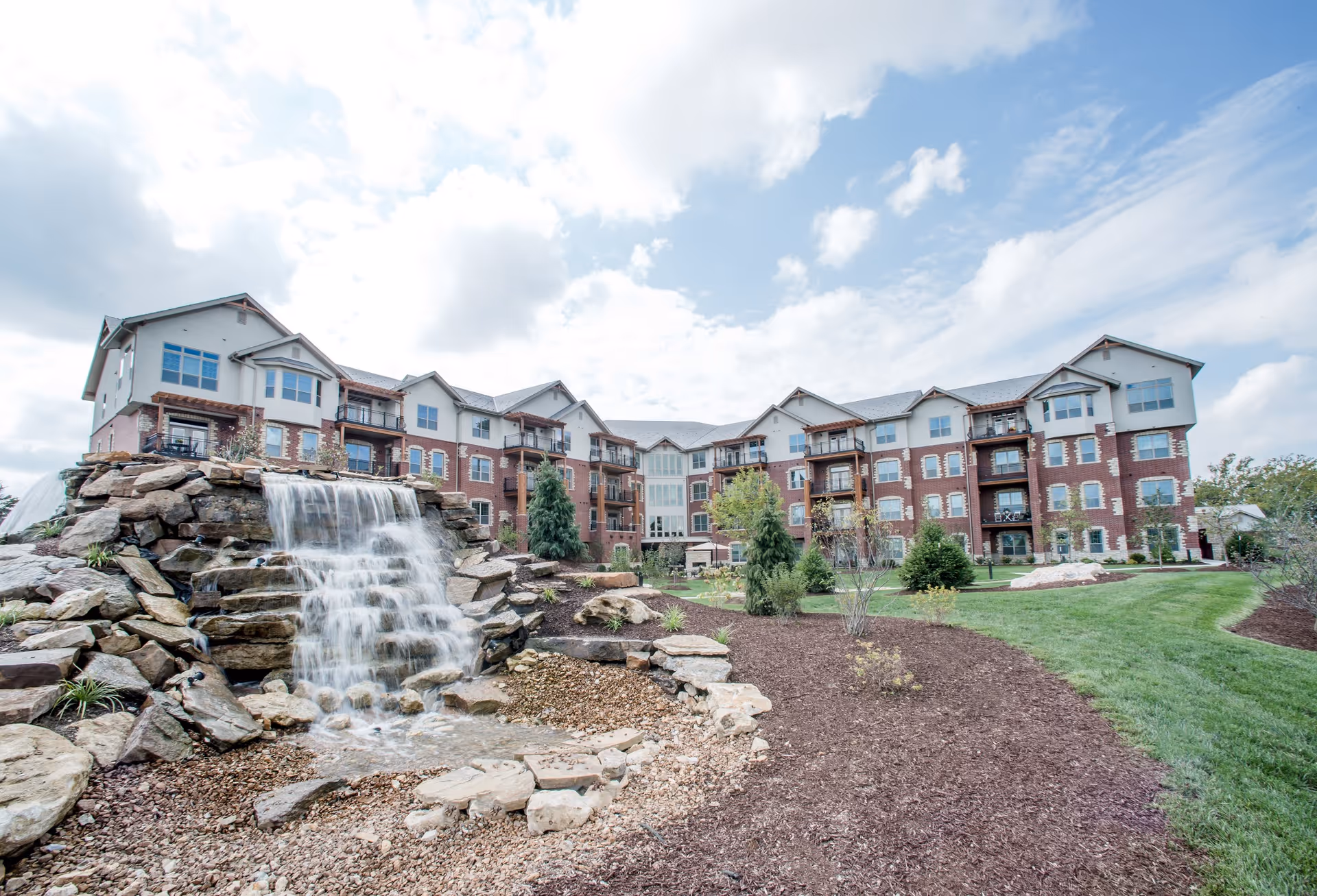 Exterior view of The Village of Bedford Walk, a multi-story senior living facility with a landscaped garden featuring a cascading waterfall over rocks, green grass, and small trees under a partly cloudy sky.