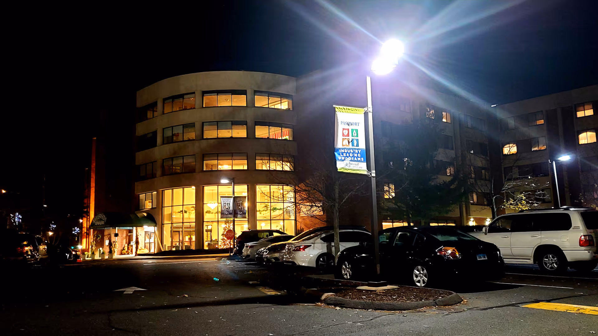 Exterior of Riverside Health and Rehabilitation Center at night, curved glass facade, street lamps, parked cars.