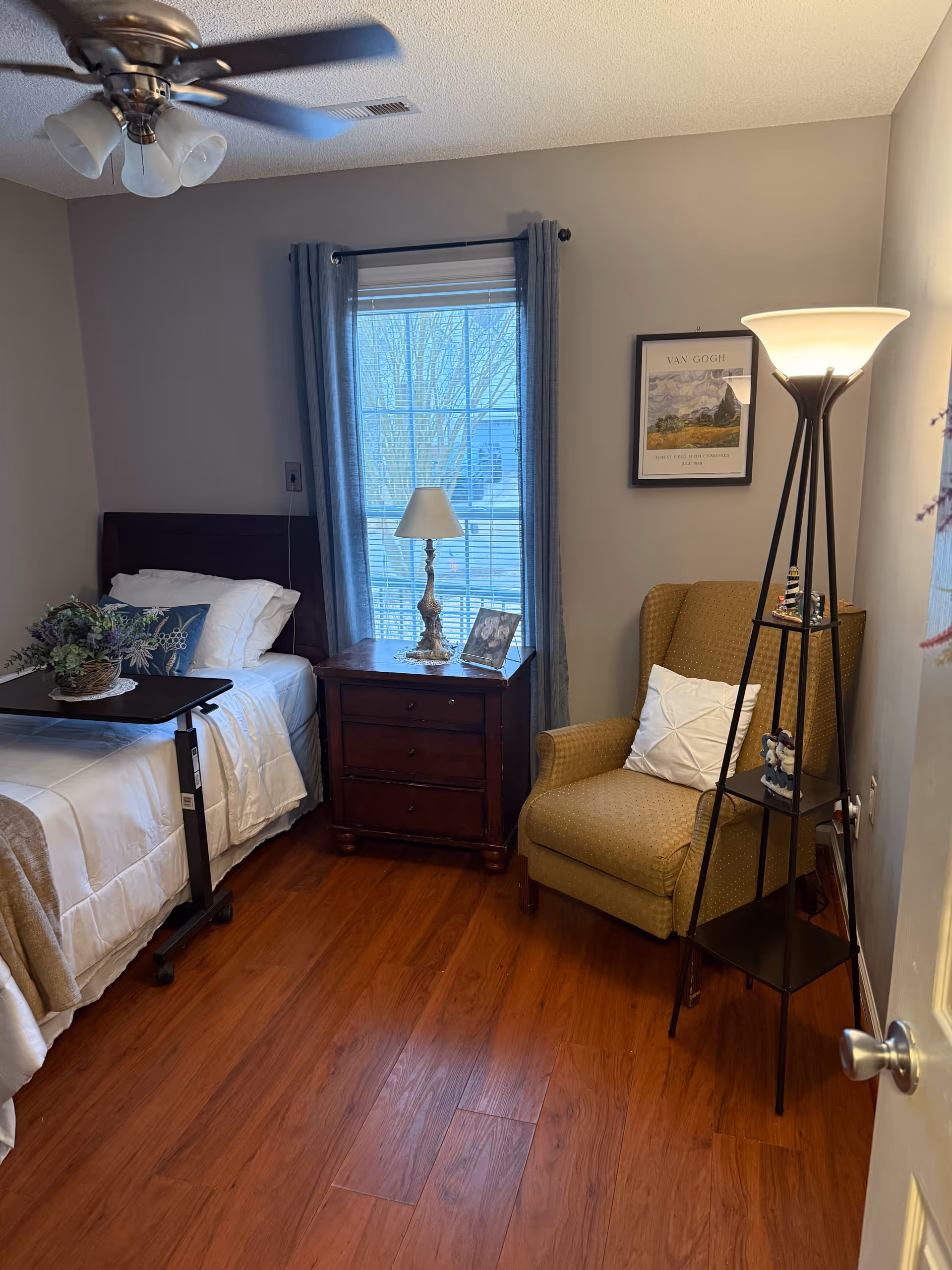 Bedroom with a bed, nightstand, window, armchair, and floor lamp in a senior living suite.