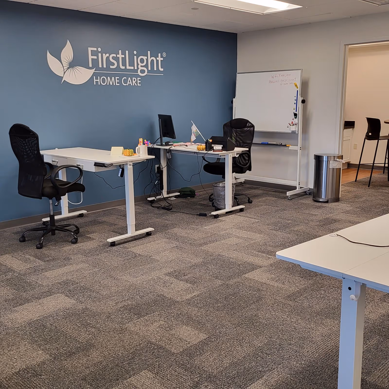 Office reception area with FirstLight Home Care logo on blue wall and desks.