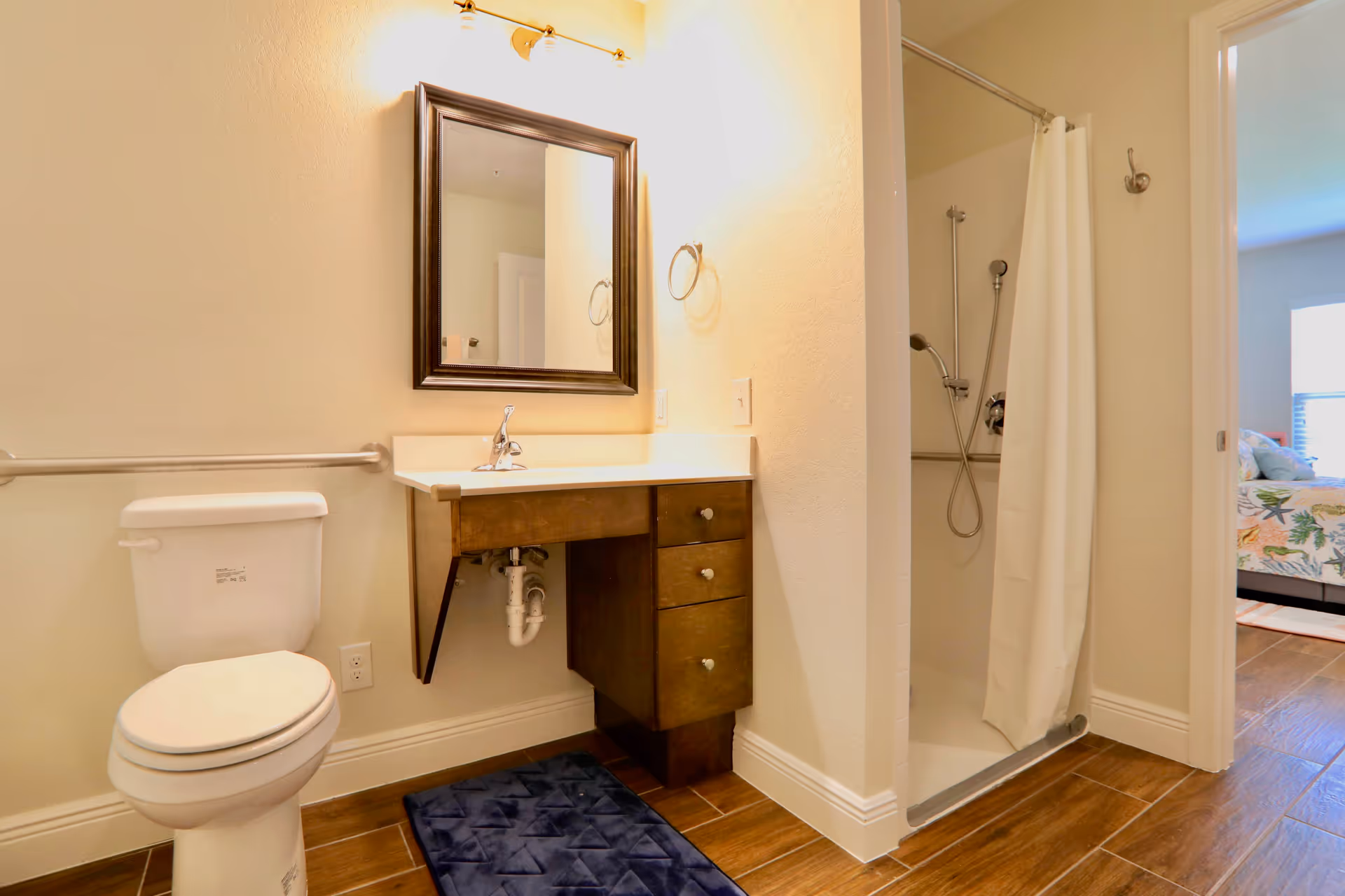 A bathroom with a toilet, a wooden vanity with a white countertop and sink, a mirror above the sink, a towel ring on the wall, and a walk-in shower with a white curtain. The bathroom floor has brown tiles, and a bedroom with a bed and window is visible through an open door.