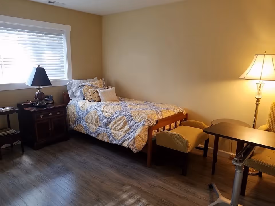 Cozy bedroom in a senior living home with bed, nightstand, lamp, and window blinds.