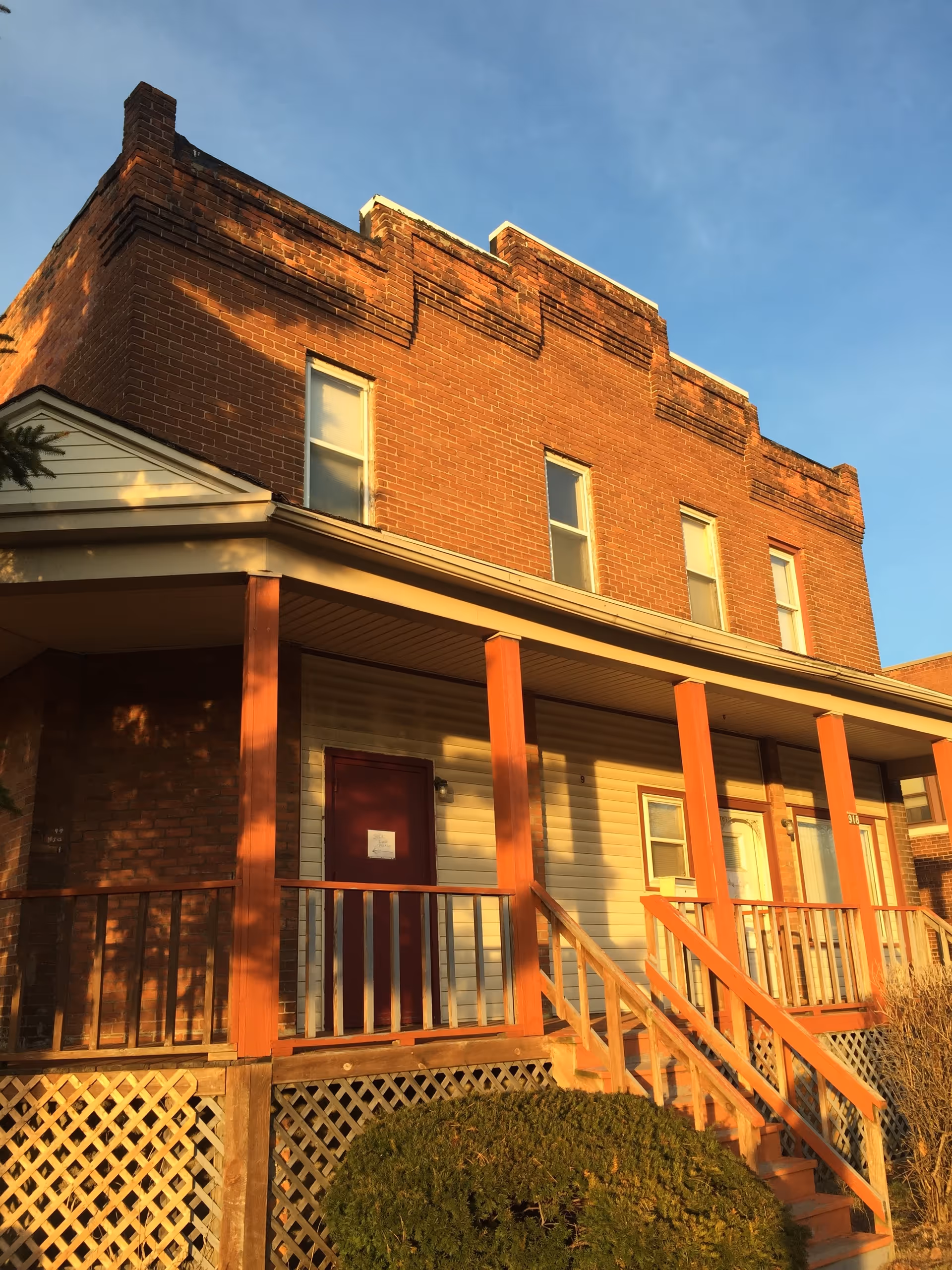 Two-story brick building with a covered porch, red posts, and wooden stair railings.