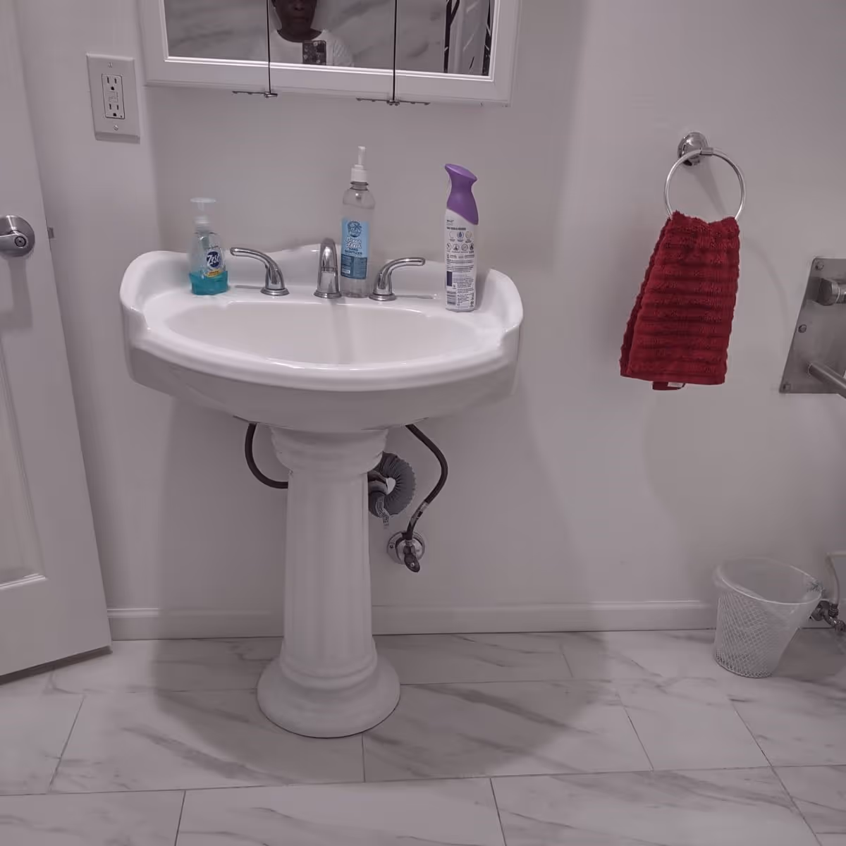 White pedestal sink with two faucets, soap bottles, and a red towel in the bathroom.
