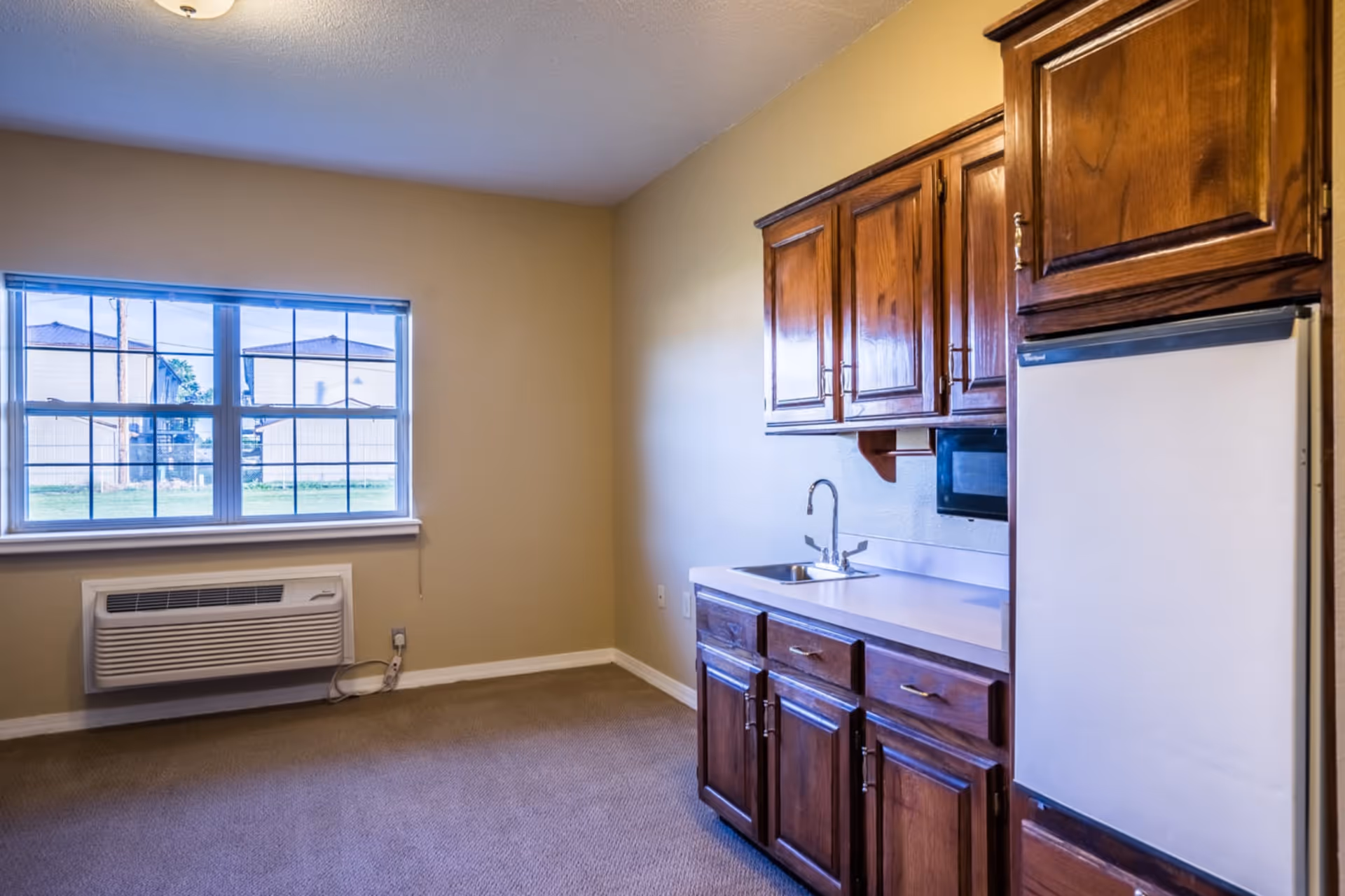 A small kitchen area with wooden cabinets, a white countertop, a sink, a microwave, and a white refrigerator. There is a window with a view of buildings outside and an air conditioning unit mounted below the window. The walls are painted beige and the floor is carpeted.
