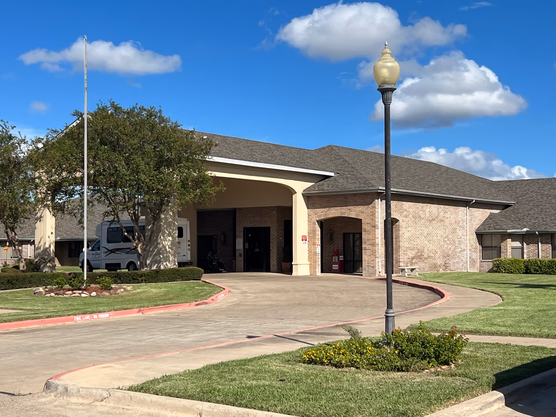 Front view of Windsor Harlingen senior living facility with brick facade, driveway, trees, and lamp post.