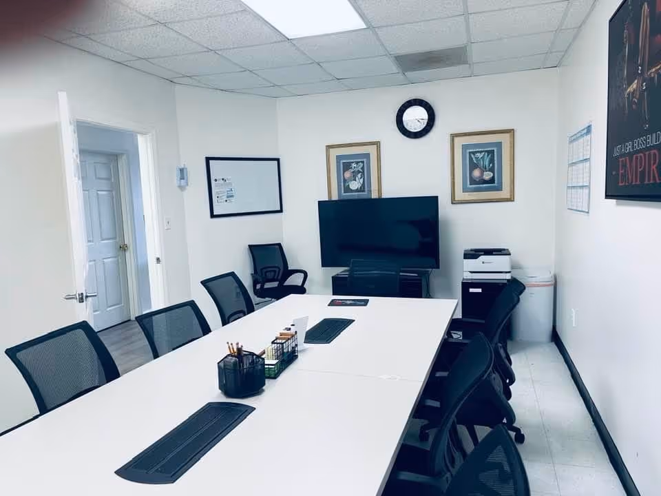 Empty conference room with a long table, mesh chairs, TV display, and printer.