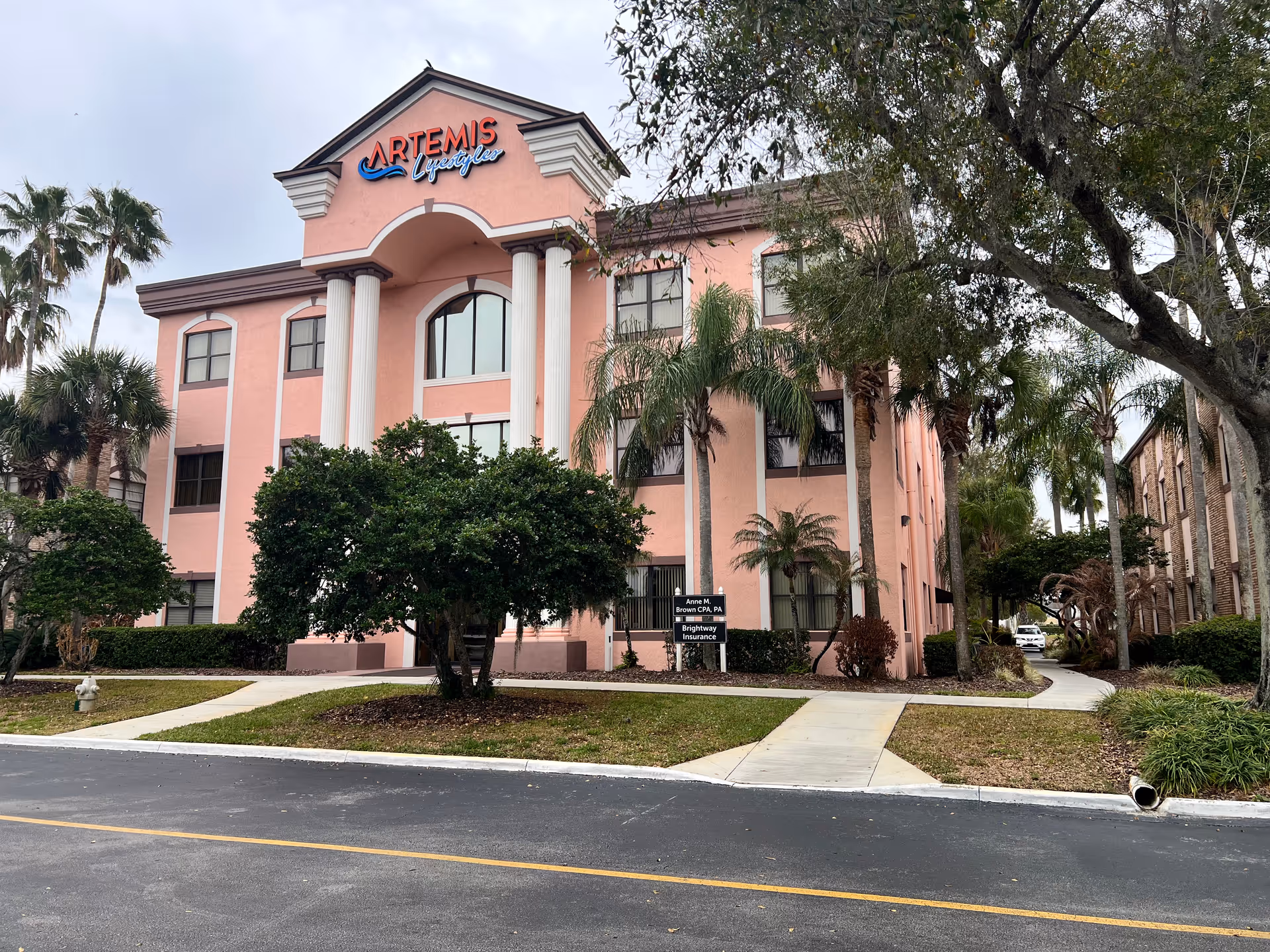 Pink three-story senior living building with white columns, palm trees, and a curved entrance.