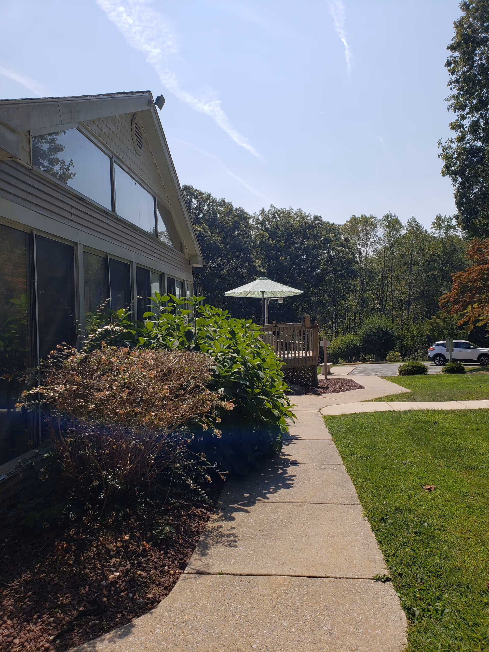 Side view of a building with large windows and a sidewalk leading to a wooden deck with a green patio umbrella. There are bushes and trees surrounding the area, with a car parked in the background under a clear sky.