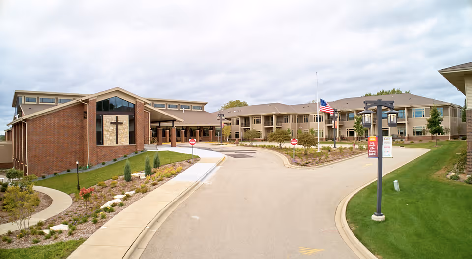 Exterior view of The Marquardt senior living facility showing a large brick building with a cross on the front, a driveway with landscaped areas, an American flag at half-mast, and a cloudy sky overhead.