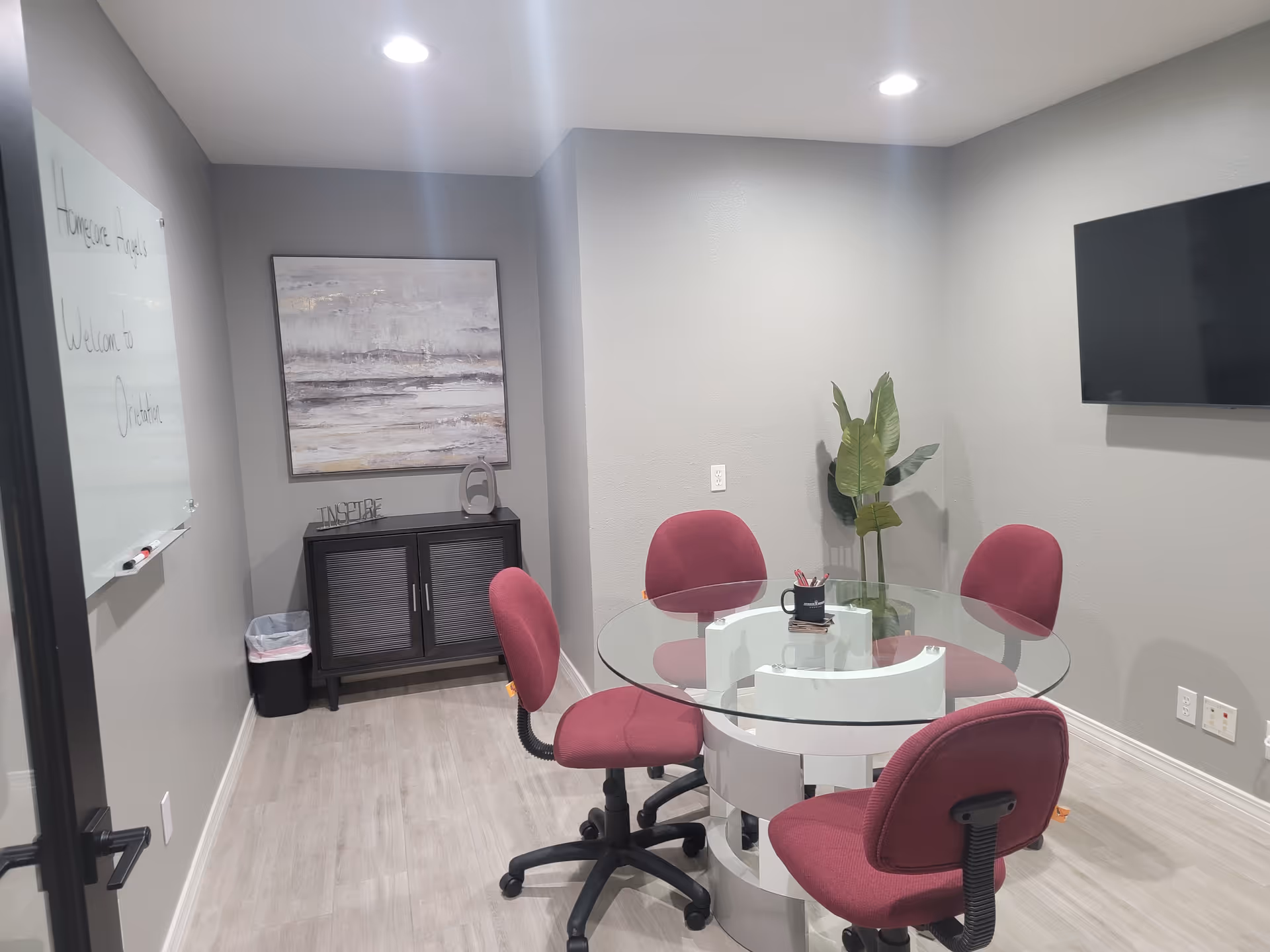 A small conference room with a round glass table, magenta chairs, a plant, and TV.